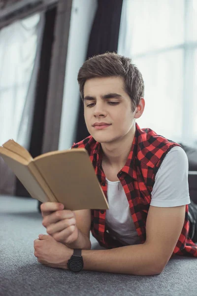 Europeo adolescente lectura libro mientras está acostado en el suelo en casa - foto de stock