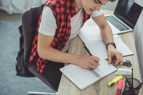 Estudante do sexo masculino estudando e escrevendo em copybooks à mesa com laptop — Fotografia de Stock