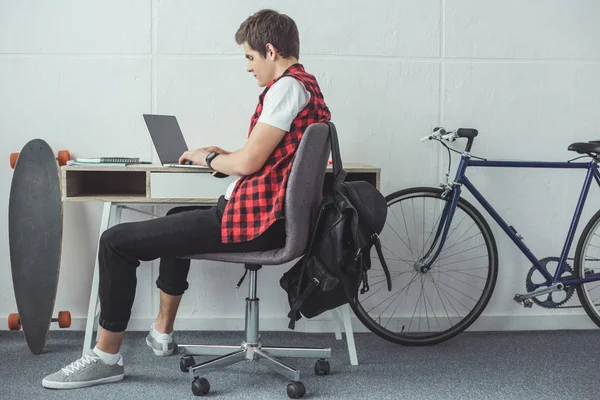 Studente che utilizza il computer portatile a tavola con skateboard e bicicletta vicino — Foto stock