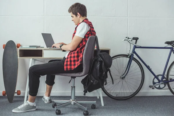 Estudiante que usa el ordenador portátil en la mesa con monopatín y bicicleta cerca — Stock Photo