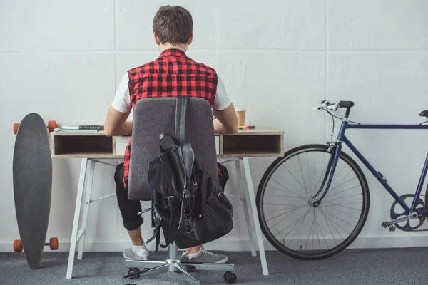 Back view of male student doing homework at table with skateboard and bicycle near — Stock Photo