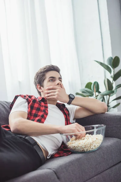 Chocado adolescente chico con palomitas de maíz viendo película en sofá en casa - foto de stock