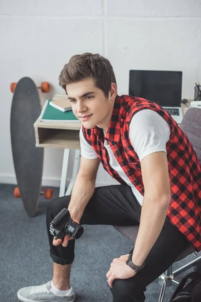 Adolescente con cámara de película retro sentado en casa con el ordenador portátil y el monopatín detrás - foto de stock