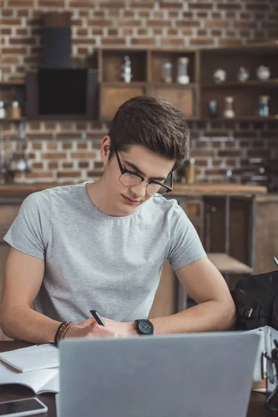 Estudiante masculino que escribe la tarea en la mesa con el ordenador portátil - foto de stock