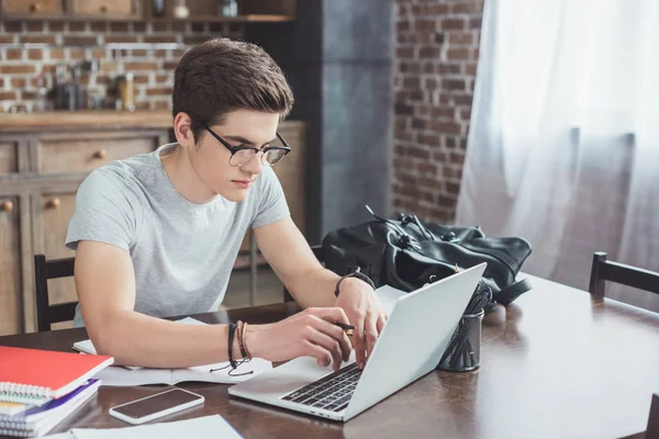 Estudiante masculino haciendo la tarea y escribiendo en el ordenador portátil en casa - foto de stock