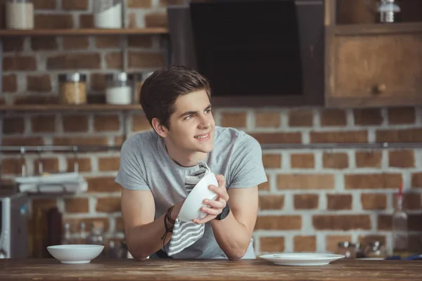 Smiling teen boy cleaning dinnerware with towel on kitchen — Stock Photo