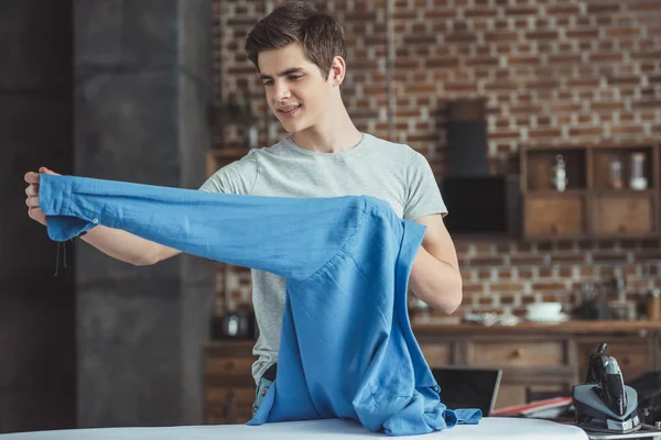 Adolescente segurando camisa azul perto de tábua de engomar com ferro — Fotografia de Stock