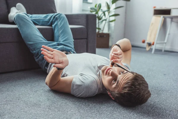 Teenager with shrug gesture talking on smartphone while lying on floor — Stock Photo