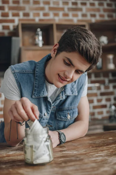 Guapo adolescente poniendo billetes de dólar en el frasco de vidrio de ahorro de dinero - foto de stock