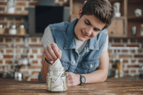 Handsome teenager taking dollar banknotes from saving glass jar for money — Stock Photo
