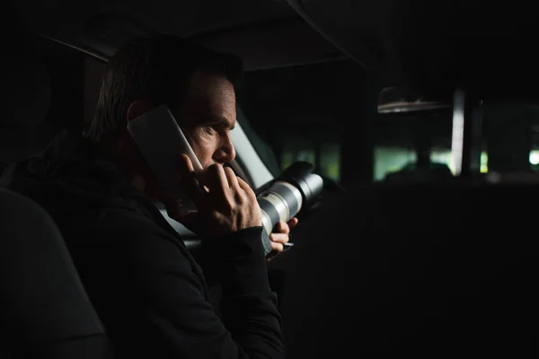 Focused male paparazzi doing surveillance by camera and talking on smartphone in car male paparazzi — Stock Photo