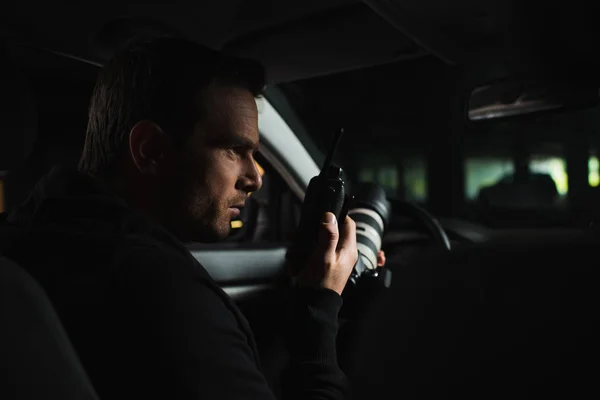 Focused male paparazzi doing surveillance by camera and using talkie walkie in car — Stock Photo