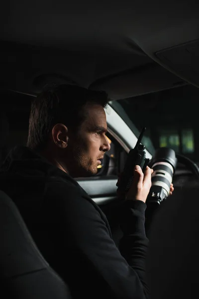Vista lateral de paparazzi macho haciendo vigilancia por cámara y usando walkie talkie en el coche - foto de stock