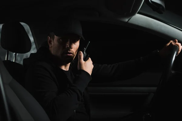 Serious man in cap doing surveillance and using talkie walkie in his car — Stock Photo