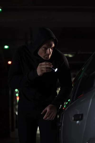 Rapinatore di sesso maschile con cappuccio nero con torcia elettrica guardando in auto — Foto stock
