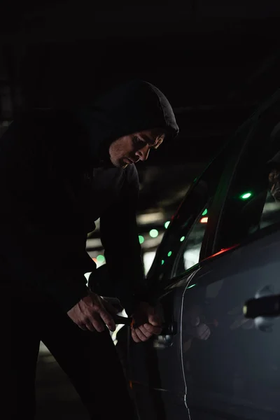 Serious male robber in black hoodie intruding car by screwdriver — Stock Photo