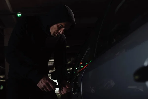 Focused male robber in black hoodie intruding car by screwdriver — Stock Photo