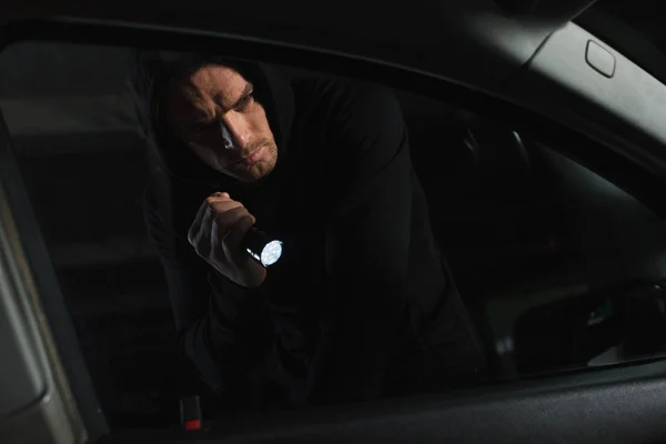 Ladrón masculino en sudadera con capucha negra con linterna mirando en el coche - foto de stock