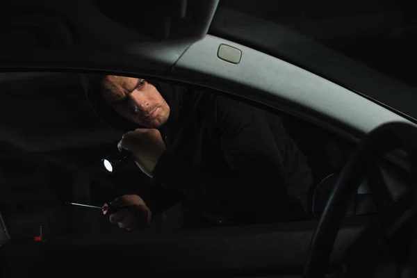 Male thief in black hoodie with flashlight and screwdriver looking into car — Stock Photo