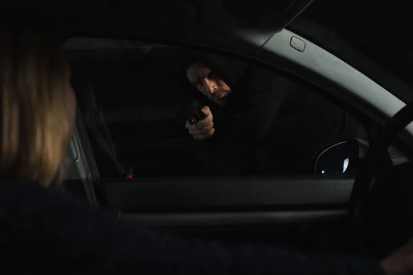 Ladrón en sudadera con capucha negra apuntando con un arma a una mujer sentada en el coche - foto de stock