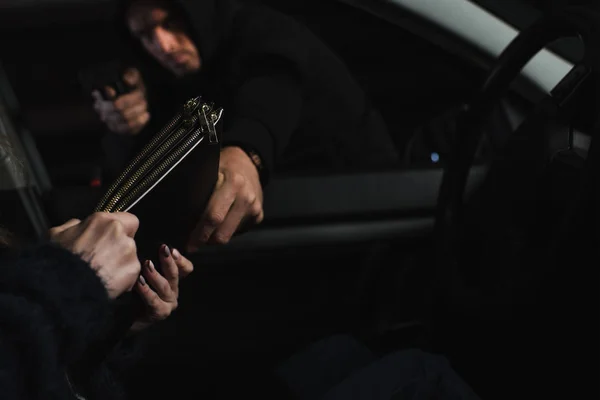 Ladrão masculino com capuz preto apontando por arma e roubando saco de mulher sentada no carro — Fotografia de Stock