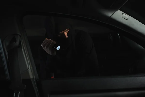 Voleur masculin en cagoule noire avec lampe de poche regardant dans la voiture — Photo de stock