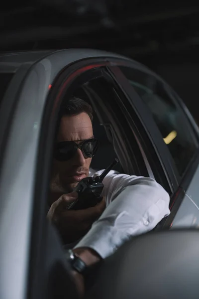 Male undercover agent in sunglasses doing surveillance and using talkie walkie in car — Stock Photo