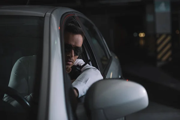 Agente encubierto masculino serio en gafas de sol haciendo vigilancia y usando walkie talkie en el coche - foto de stock