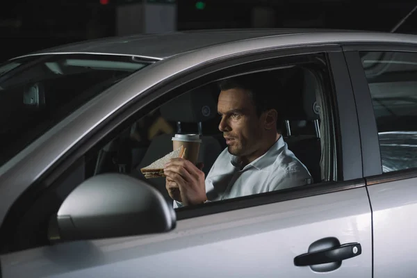Seitenansicht eines männlichen Privatdetektivs, der Kaffee trinkt und im Auto Sandwich isst — Stockfoto