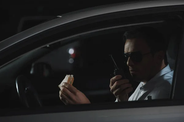 Male undercover agent in sunglasses having lunch and using talkie walkie in car — Stock Photo