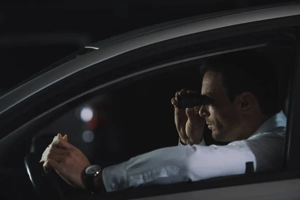 Vista lateral del agente encubierto haciendo vigilancia por prismáticos y comiendo sándwich en el coche - foto de stock