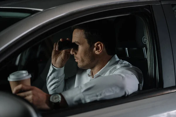 Side view of undercover male agent doing surveillance by binoculars and drinking coffee in car — Stock Photo