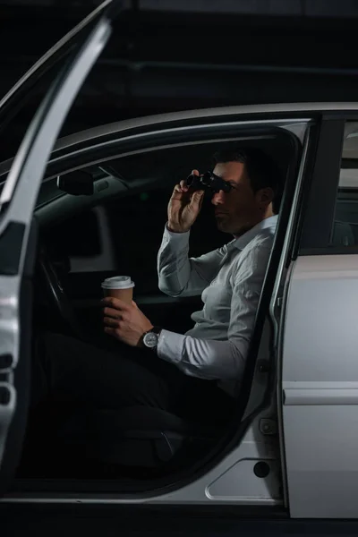 Undercover male agent spying by binoculars and drinking coffee in car — Stock Photo