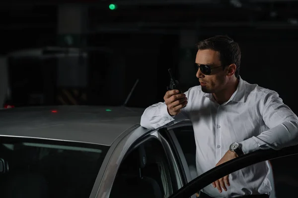 Agente disfarçado masculino em óculos de sol usando walkie talkie perto do carro — Fotografia de Stock