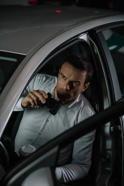 Vista de ángulo alto de detective privado masculino con taza de café haciendo vigilancia por prismáticos desde el coche - foto de stock