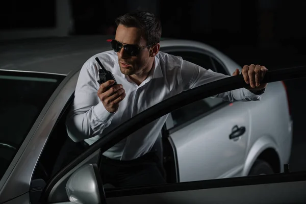 Agente encubierto masculino serio en gafas de sol usando walkie talkie cerca del coche - foto de stock