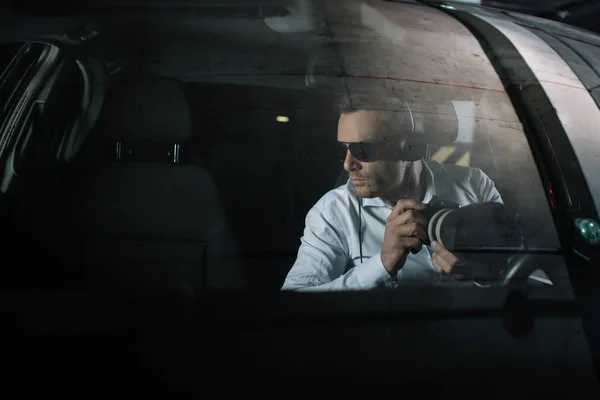 Serious male private detective in headphones doing surveillance by camera with object glass from car — Stock Photo