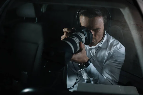 Focused male private detective in headphones doing surveillance by camera from car — Stock Photo