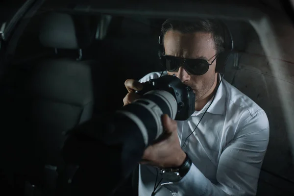 Male private detective in headphones doing surveillance by camera with object glass from car — Stock Photo