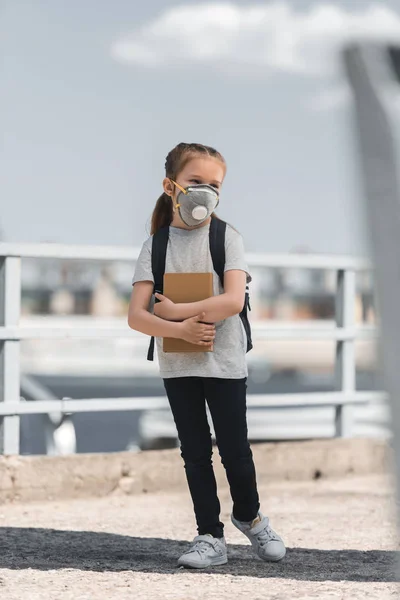 Niño en máscara protectora de pie con bolsa y libro sobre puente, concepto de contaminación del aire — Stock Photo