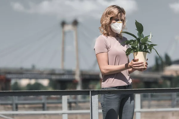 Woman in protective mask holding potted plant on street, air pollution concept — Stock Photo