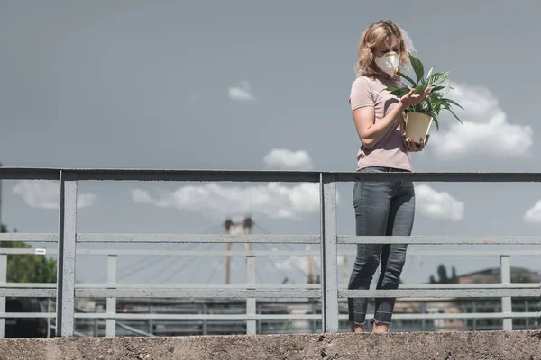 Woman in protective mask looking at leaves of potted plant on bridge, air pollution concept — Stock Photo