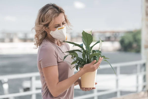 Mulher em máscara protetora olhando para planta em vaso na ponte, conceito de poluição do ar — Fotografia de Stock