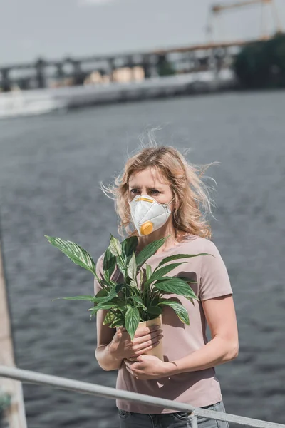 Femme en masque de protection tenant une plante en pot près de la rivière, concept de pollution atmosphérique — Photo de stock