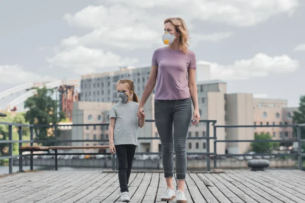 Mother and daughter in protective masks holding hands on pier and looking away, air pollution concept — Stock Photo