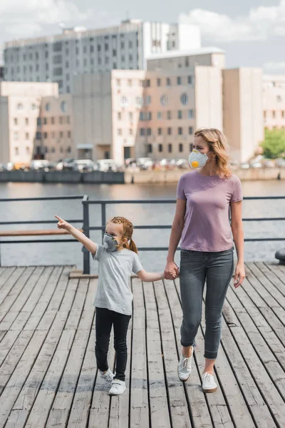 Madre e hija en máscaras protectoras cogidas de la mano en el muelle cerca del río, concepto de contaminación del aire - foto de stock