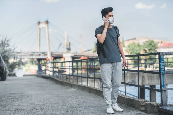 Asian teen in protective mask talking by smartphone on bridge, air pollution concept — Stock Photo