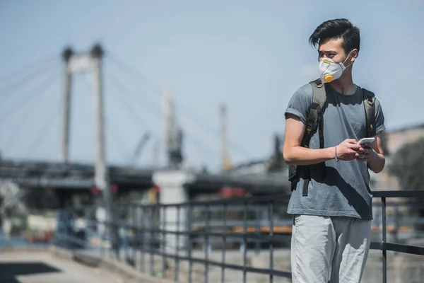 Asiático adolescente en protección máscara celebración smartphone y mirando lejos, aire contaminación concepto - foto de stock