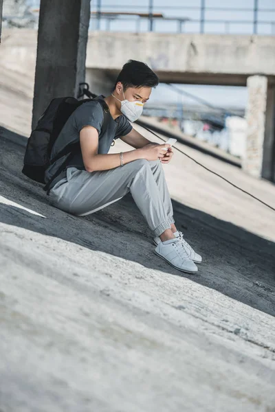 Vista lateral de asiático adolescente em máscara protetora usando smartphone sob ponte, conceito de poluição do ar — Fotografia de Stock