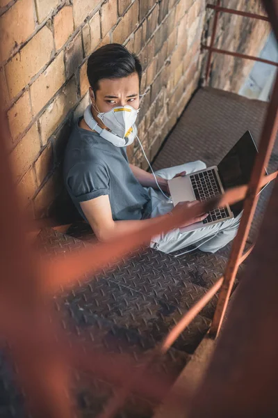 Високий кут зору азіатського підлітка в захисній масці, що сидить з ноутбуком на сходах, концепція забруднення повітря — стокове фото